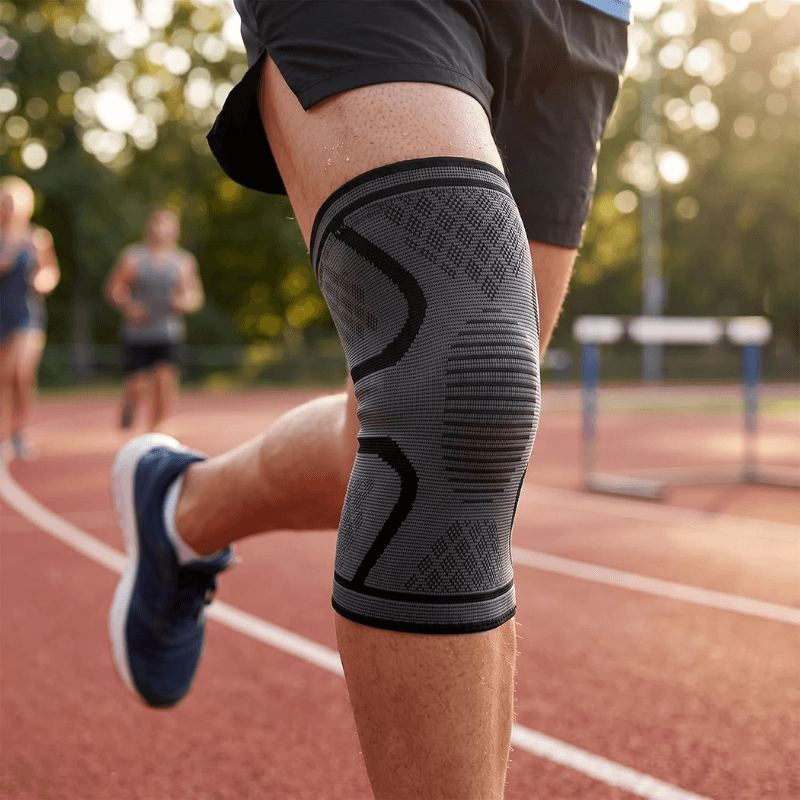 A close-up of a person's leg on a running track wearing a gray and black knee compression sleeve, with blurred runners and hurdles in the background.